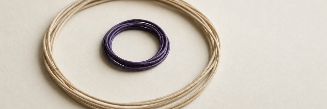 Two loops of waxed cord on pale paper: a small tight violet coil lying inside a much larger loose loop of undyed cord.